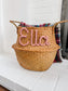Image of a seagrass brown basket with "Ella" written in dusty pink colored yarn, containing a cozy throw blanket, placed on a living room fireplace.